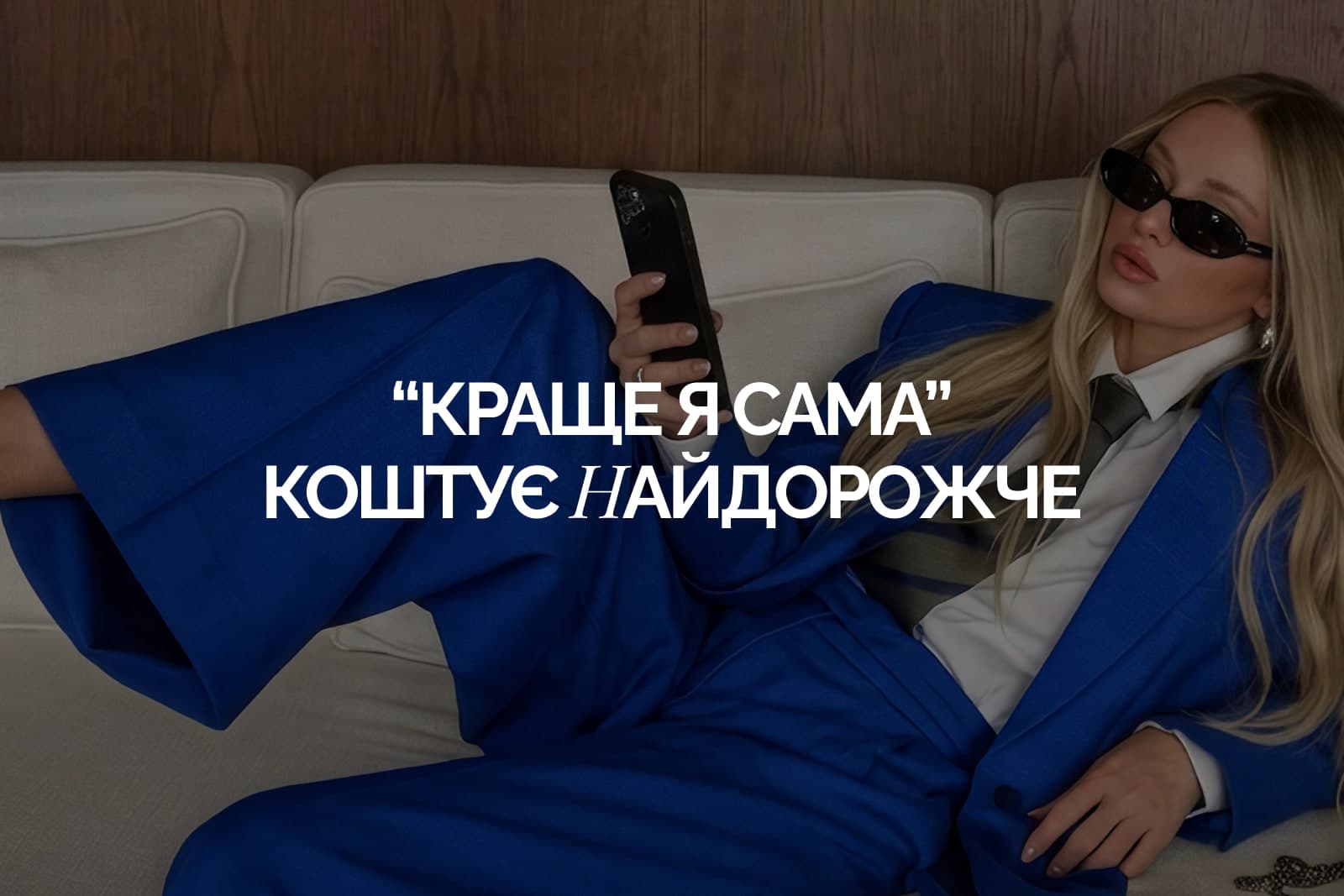 Жінка в синьому костюмі й темних окулярах лежить на дивані з телефоном у руці